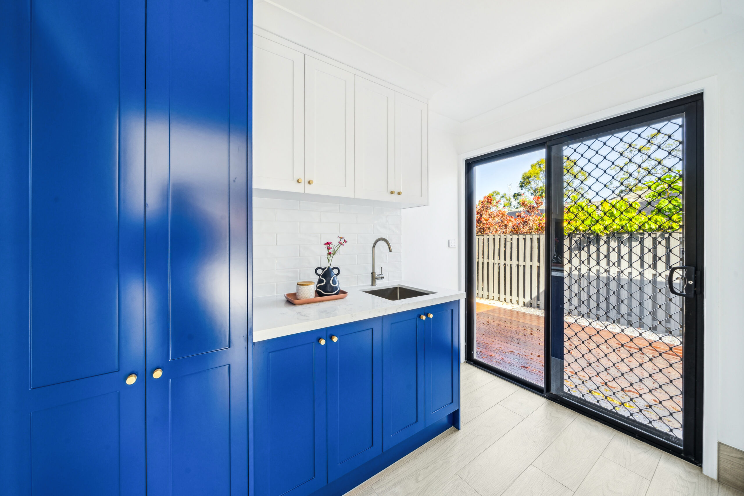 Renovated laundry with blue shaker cabinetry, floor to ceiling storage, stone benchtop and a sliding door to the deck